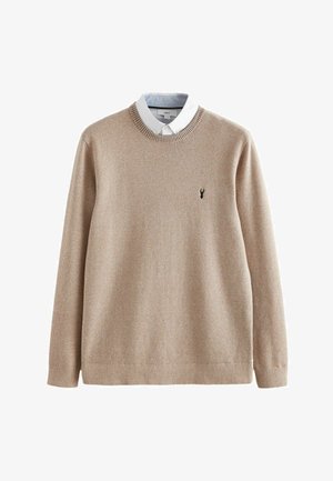 Beige gestrickter Pullover mit geripptem Muster, Rundhalsausschnitt und einem weißen Hemd darunter. Kleines schwarzes Logo auf der Brust.