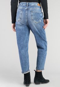 Jean en denim bleu clair avec une coupe décontractée, présentant des ourlets roulés et deux poches arrières avec un motif de couture distinct. Associé à des bottines noires.