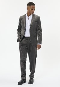 Pelēks samta uzvalks ar vienrinds jaku un saskanīgiem biksēm. Zemāk balta krekls, apvienots ar melnām kurpēm, minimāls aksesuārs.