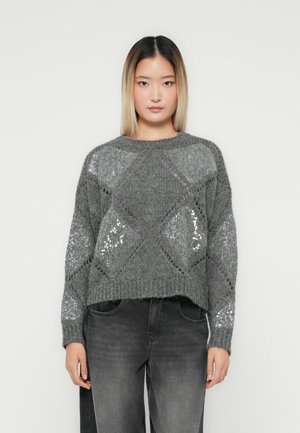 Vrouw met steil, schouderlang donkerblond haar draagt een grijze gebreide trui met een patroon van pailletten in diamanten en vervaagde zwarte jeans.