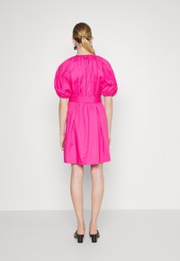 Woman with blonde ponytail wearing a short-sleeve, bright pink dress with a cinched waist and black heeled sandals, standing against a white background.