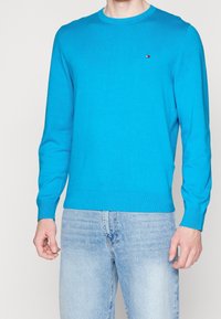 Klart blå, långärmad stickad tröja med rund halsringning och ribbade muddarna. Har en liten logotyp på bröstet. Matchas med ljusa denimjeans.