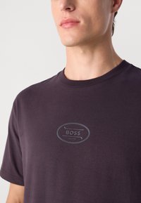 Közelkép egy személyről, aki sötétlila pólót visel, amelyen a mellkason egy kis ovális "BOSS EST. 1927" logó található.