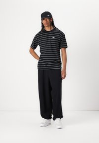 Schwarz-weiß gestreiftes T-Shirt mit kurzen Ärmeln, schwarze Hose, schwarze Kappe mit Logo und weiße Sportschuhe. Lässige Streetwear-Ästhetik.