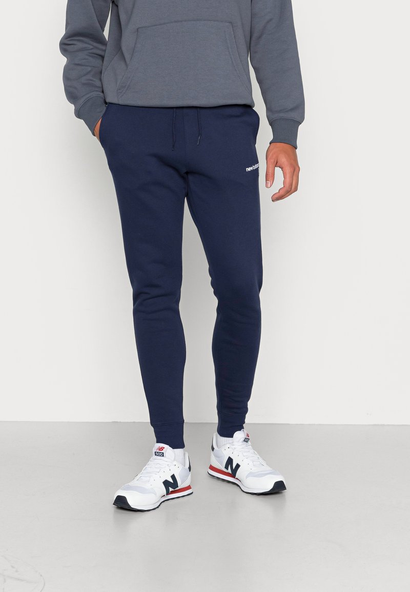 Pantalons de survêtement navy avec taille à cordon, poignets côtelés et accent de logo blanc, associés à un sweat à capuche gris et des baskets blanches.