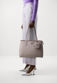 Sac fourre-tout en cuir beige avec quincaillerie argentée, doté d'une fermeture éclair et d'une pochette amovible. Associé à des talons argentés et un pantalon violet clair.