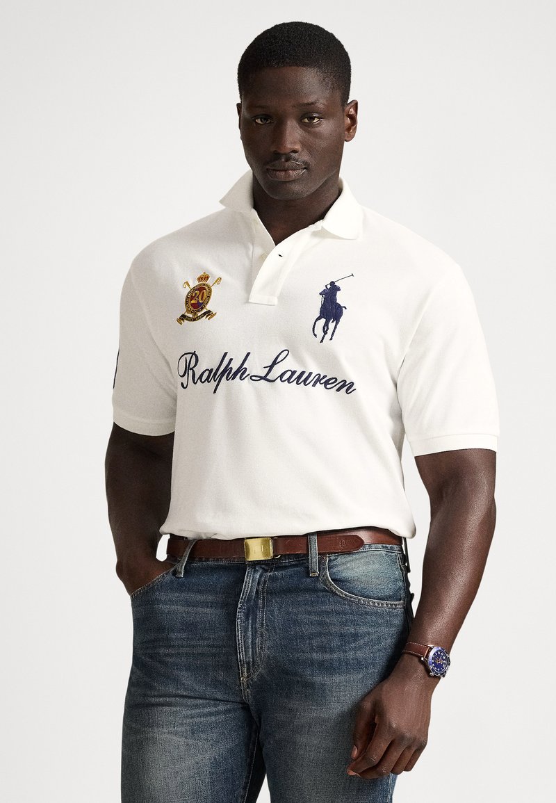 Moški v beli Ralph Lauren polo majici z vezenimi logotipi, modrih kavbojkah, rjavem usnjenem pasu in uri z rjavim usnjenim paščkom.