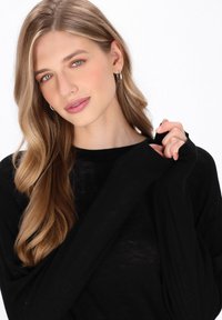Maglia nera a maniche lunghe in maglia con vestibilità rilassata, caratterizzata da polsini a costine e una texture morbida. Modella che tiene il tessuto vicino al colletto.