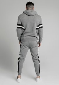 Mężczyzna z tatuażem na szyi, ubrany w szary dres w czarno-białe paski, stojący plecami na jednolitym tle.