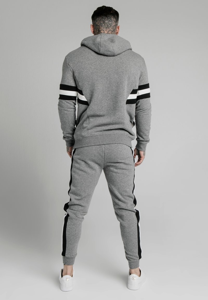 Mężczyzna z tatuażem na szyi, ubrany w szary dres w czarno-białe paski, stojący plecami na jednolitym tle.