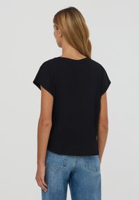 T-shirt nera in cotone con vestibilità ampia, maniche corte, scollatura rotonda e orlo corto, indossata con jeans blu a vita alta.