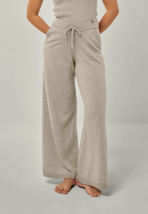 Frau trägt beigen weiten Strickhose mit Fronttaschen und Kordelzugbund, steht barfuß vor einem neutralen Hintergrund.