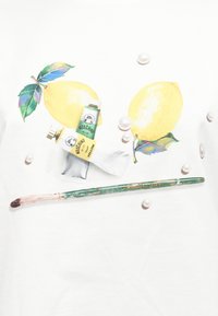 Δύο κίτρινα λεμόνια με πράσινα φύλλα, σωληνάρια χρώματος με την ετικέτα "Moschino" σε κίτρινο και πράσινο, σκορπιστές πέρλες και ένα πράσινο πινέλο πάνω σε λευκό φόντο.