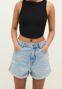Nainen, jonka yllä on vaaleansiniset korkeavyötäröiset farkkushortsit, joissa on käänteet, ja musta hihaton cropped-top, kädet taskuissa, neutraali tausta.