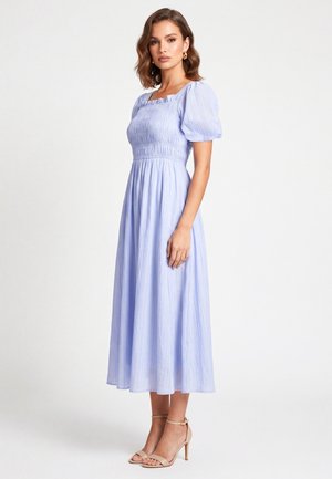 Vrouw draagt een lichtblauwe midijurk met pofmouwen en een geplooid lijfje, gecombineerd met beige enkellaarsjes met bandje, staand tegen een witte achtergrond.