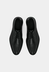 Um par de sapatos sociais pretos de couro com detalhes em brogue e atacadores, vistos de cima sobre um fundo branco.