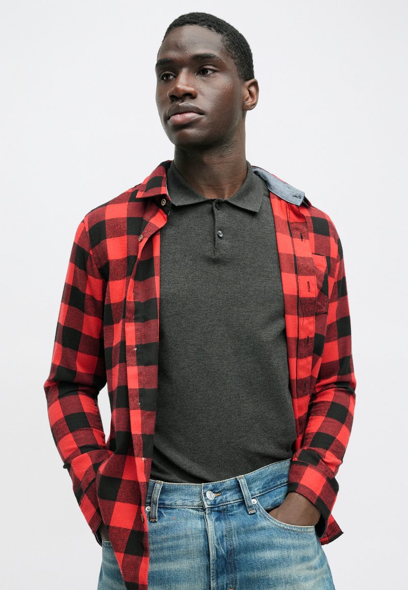 Red and black plaid flannel shirt layered over a dark gray polo, worn with light blue denim jeans. Features button details and contrasting collar.