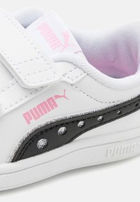 Weißer Sneaker mit schwarzen Akzenten, Glitzerdetails und pinkem Branding. Verfügt über einen Klettverschluss und ein strukturiertes Stofffutter.