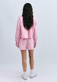 Rosa Kurzarmhemd mit kastiger Schnitt, kombiniert mit passenden Shorts. Der Stoff wirkt glatt; es hat lange Ärmel und geknöpfte Manschetten. Weiße Sneakers.