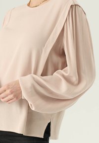Jasnoróżowa bluzka z fakturowanej tkaniny, luźny krój i bufiaste rękawy. Posiada boczne rozcięcia i okrągły dekolt.