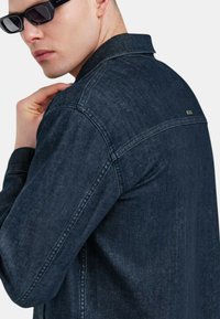 Giacca di jeans blu scuro con colletto, chiusura frontale a bottoni e cuciture a vista. Presenta un piccolo etichetta del logo sul retro vicino al collo.