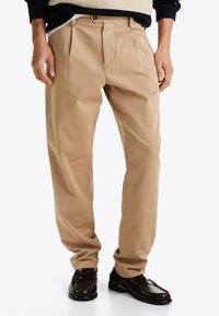 Beige plissierte Hose mit glatter Textur, mit einem Knopfverschluss und Seitentaschen, kombiniert mit dunklen Lederschuhen.