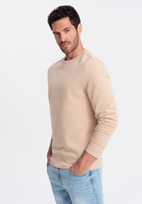 Sudadera beige hecha de una tela suave, con cuello redondo y puños acanalados. Combinada con jeans de color azul claro para un look casual.