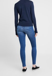 Kvinna i en marinblå långärmad tröja, blå slim fit jeans och svarta låga sneakers, stående med ryggen mot kameran.