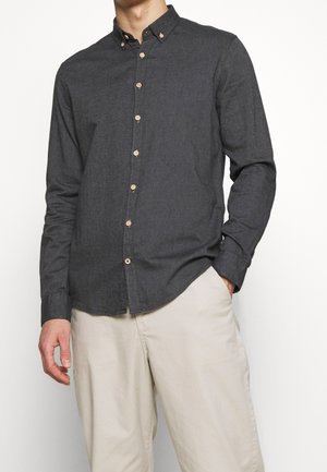 Mężczyzna w ciemnoszarej koszuli na guziki i beżowych spodniach, z jedną ręką w kieszeni, stojący na tle jednolitego białego tła.