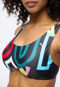 Γυναίκα που φοράει μια πολύχρωμη αθλητική σουτιέν με αφηρημένα γεωμετρικά μοτίβα και το λογότυπο της Puma στο αριστερό μέρος, πάνω σε απλό φόντο.