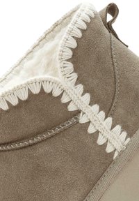 Botín de ante taupe con forro de lana, costura blanca decorativa y suela de goma texturizada. Cuenta con un lazo para facilitar el uso.