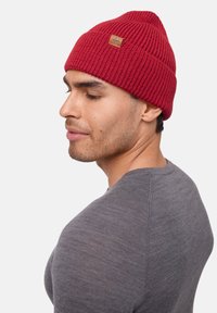 Red knit beanie with a ribbed texture and pointed top, featuring a small leather tag. Worn with a gray fitted sweater.