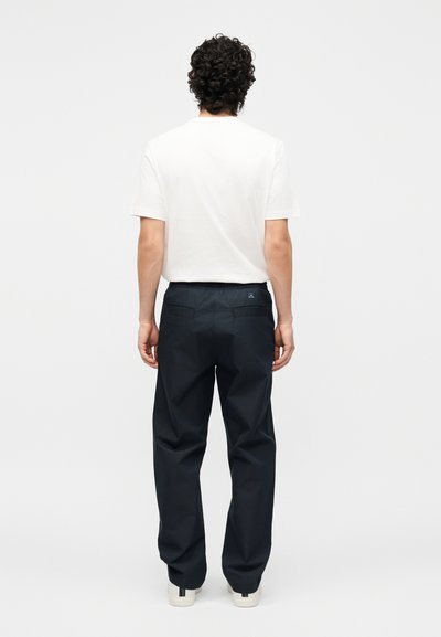 Άνδρας με σγουρά σκουρόχρωμα μαλλιά, ντυμένος με λευκό κοντομάνικο πουκάμισο και μαύρο παντελόνι, στέκεται με την πλάτη γυρισμένη σε ένα απλό λευκό φόντο.