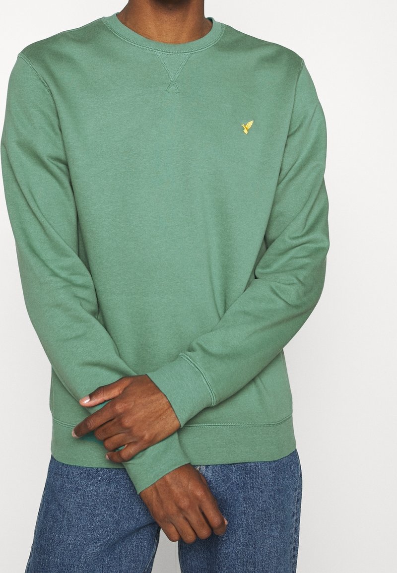Sudadera verde hecha de una mezcla de algodón, con cuello redondo y puños acanalados. Un pequeño logotipo amarillo es visible en el pecho.