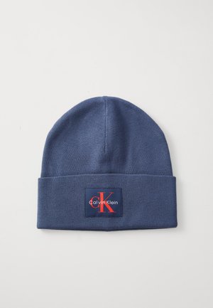 Tumesinine kootud beanie, millel on volditud äär, ja millel on punase ja valge neljakujuline logoipatch, kus on kirjas "Calvin Klein" ja "CK."