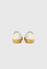 Light beige canvas shoes with a low top, rubber sole, and rounded toe. Features a small logo detail on the heel and a textured surface.