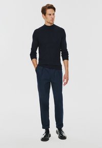 Granatowy sweter z prążkowanym golfem i dobrze skrojone spodnie w tym samym kolorze. Model stoi w czarnych sznurowanych butach, z rękami w kieszeniach, minimalistyczny design.