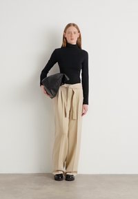 Crna majica s visokim izrezom, krem hlače širokih nogavica s vezanom strukom, crna torba od mekane kože i masivne crne cipele. Jednostavna pozadina.