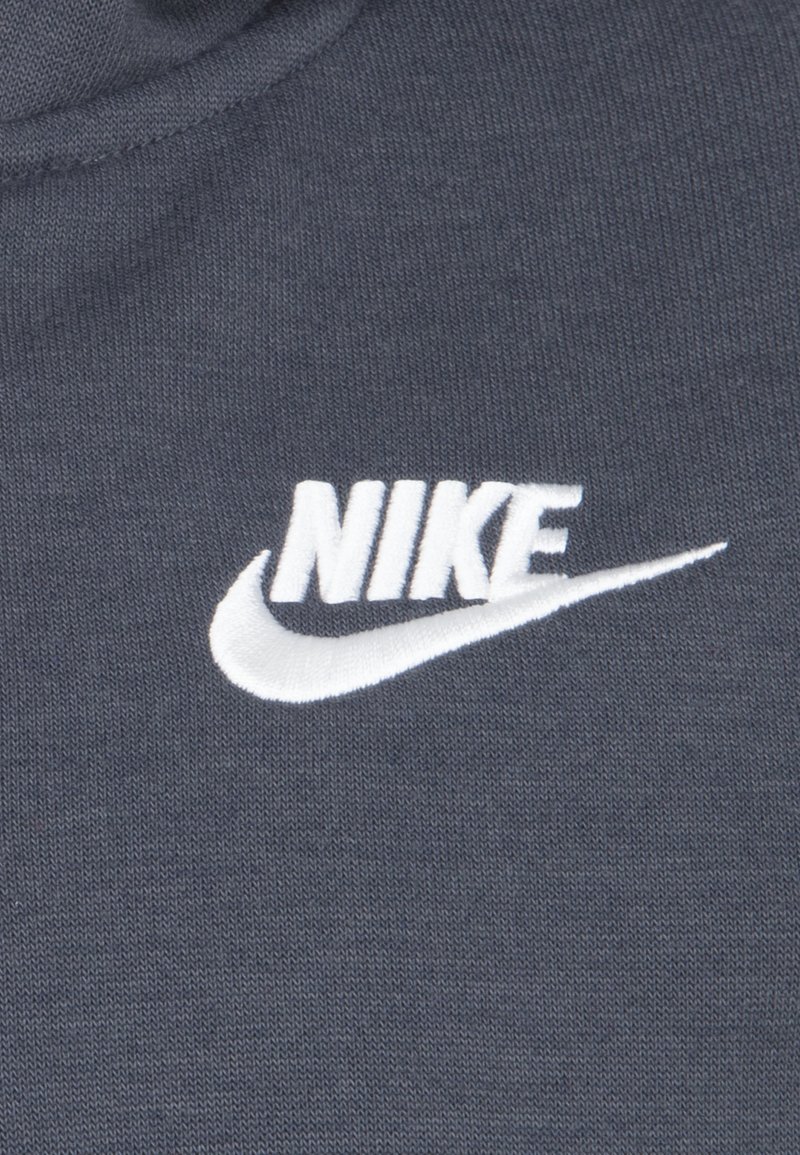 Un tissu en coton gris présente un logo Nike brodé blanc en évidence, mettant en lumière un design en forme de swoosh. La texture est lisse et uniforme.