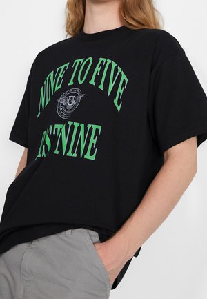 T-shirt noir avec un texte vert indiquant "NINE TO FIVE" et "PST NINE" porté par une personne avec la main dans la poche d'un pantalon gris.