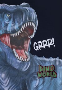 T-shirt con un'illustrazione di un Tirannosaurus rex blu e grigio con denti affilati e bocca aperta. Testo "GRRR!" e "DINO WORLD" in stile grafico.