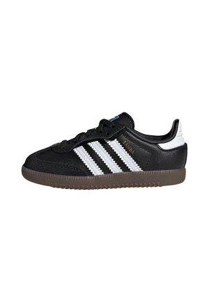 Zapatilla negra Adidas Samba con rayas blancas, suela marrón y lengüeta blanca en el talón, mostrada en perfil lateral derecho.