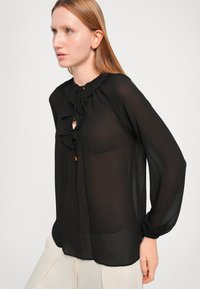 Svart bluse i gjennomsiktig stoff med en volangkant ved halsen, knyte-detail og lange ermer med elastiske mansjetter. Glatt tekstur, lett design.
