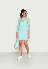 Um vestido verde menta justo com uma sobreposição longa de mangas transparentes em padrão mármore. Os acessórios incluem uma bolsa de ombro azul clara e ténis brancos.