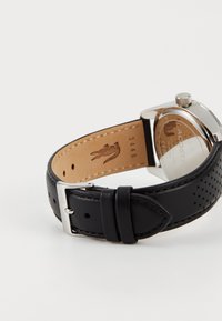 Relógio com pulseira de couro preto com perfurações, caixa de aço inoxidável prateado e mostrador redondo e transparente. Apresenta um forro interior castanho com logotipo.