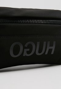 Pochette en tissu noir avec une fermeture éclair horizontale visible et le texte inversé "HUGO" imprimé en gris foncé.