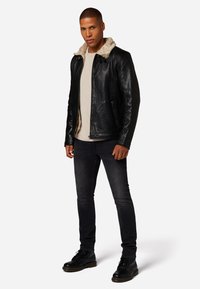 Zwarte leren jas met een bontgevoering kraag, gedragen over een lichtgekleurde shirt, gecombineerd met donkere jeans en zwarte boots, rechtopstaand.