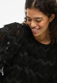 Haut noir texturé avec des motifs ondulés et des détails en forme de plumes sur les manches. Encolure ronde et tissu léger et transparent.