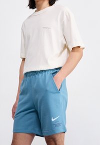 Ljusbeige bomulls-t-shirt med korta ärmar och subtil text på framsidan, tillsammans med blå Nike sportshorts med sidfickor.