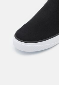 Vue avant d'une chaussure noire en tissu à enfiler avec une semelle en caoutchouc blanche texturée et un bord noir autour du bord de la semelle.